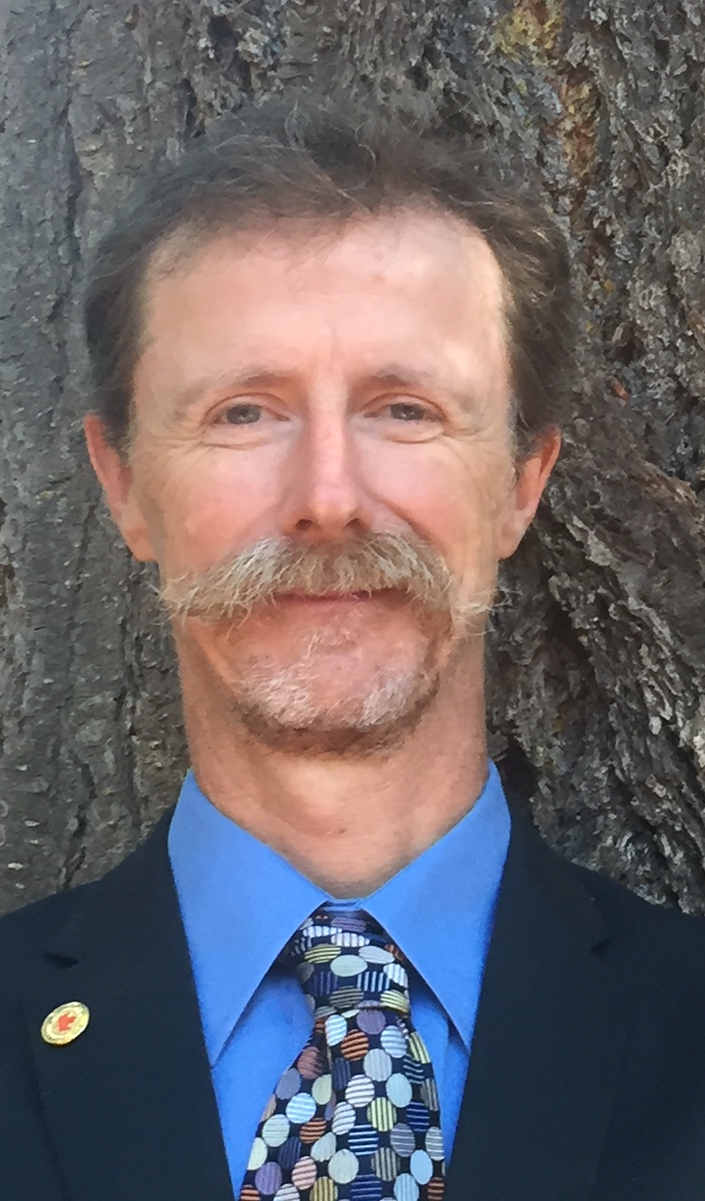 Un homme blanc mince d'un certain âge avec une moustache observe la caméra. Il porte un costume noir et une chemise bleue ainsi qu'une cravate.