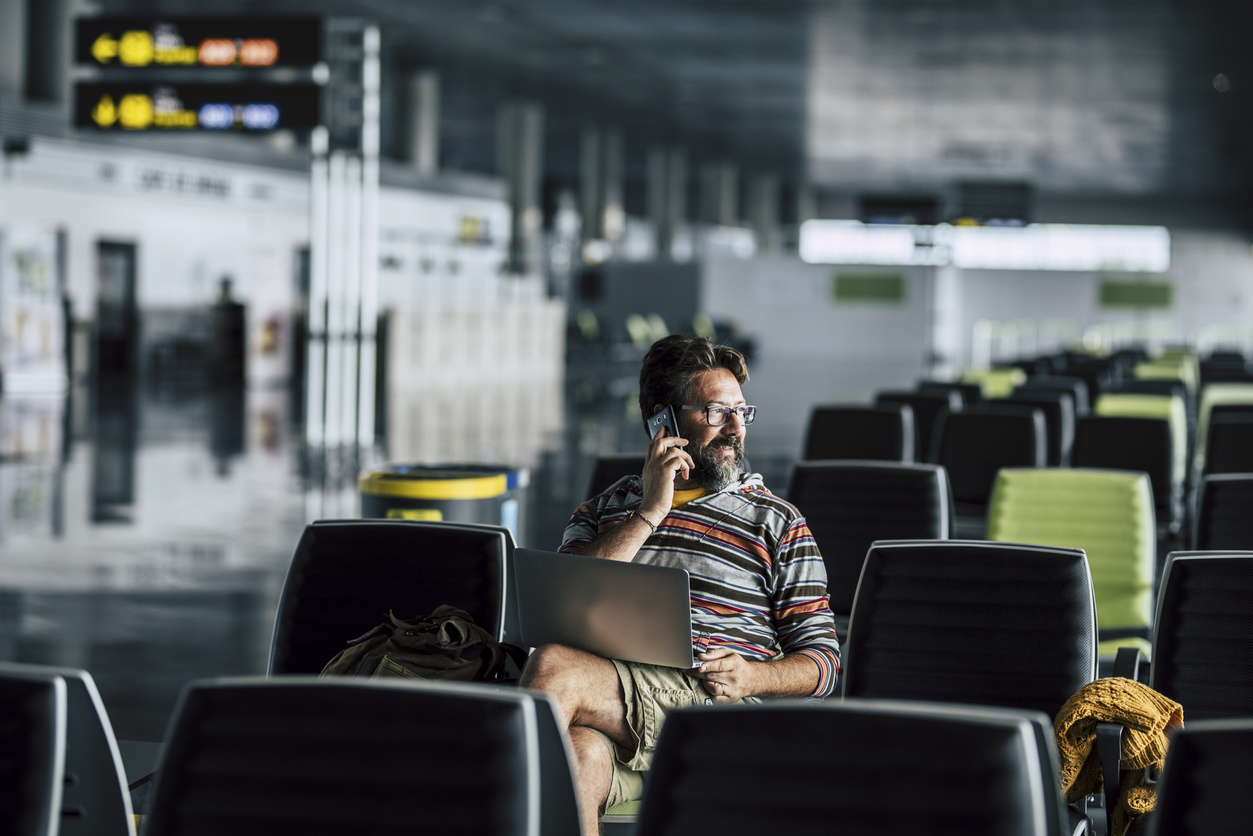 Homme seul assis et utilisant un appareil technologique comme un ordinateur portable et un téléphone mobile à la porte de l'aéroport en attendant son vol