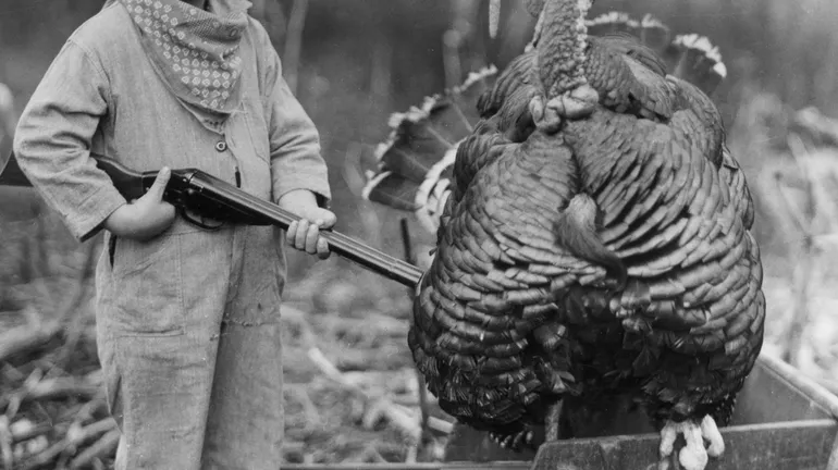 Vers 1935 : Photo en pied d’un jeune garçon souriant, debout à l’extérieur près d’une dinde sauvage, un fusil à la main.