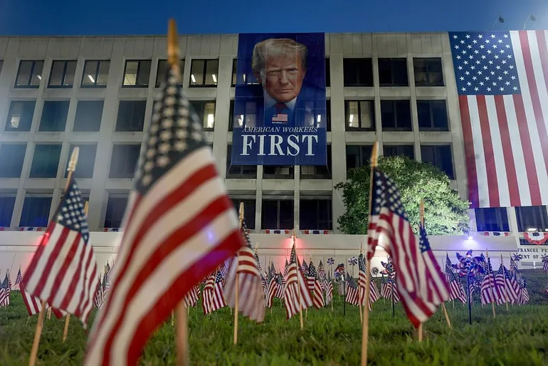 Des drapeaux américains sont plantés dans le sol devant une banderole à l’effigie du président américain Donald Trump, accrochée au bâtiment du ministère américain du Travail, en prévision des festivités du 4 juillet, le 3 juillet 2026, à Washington, DC.